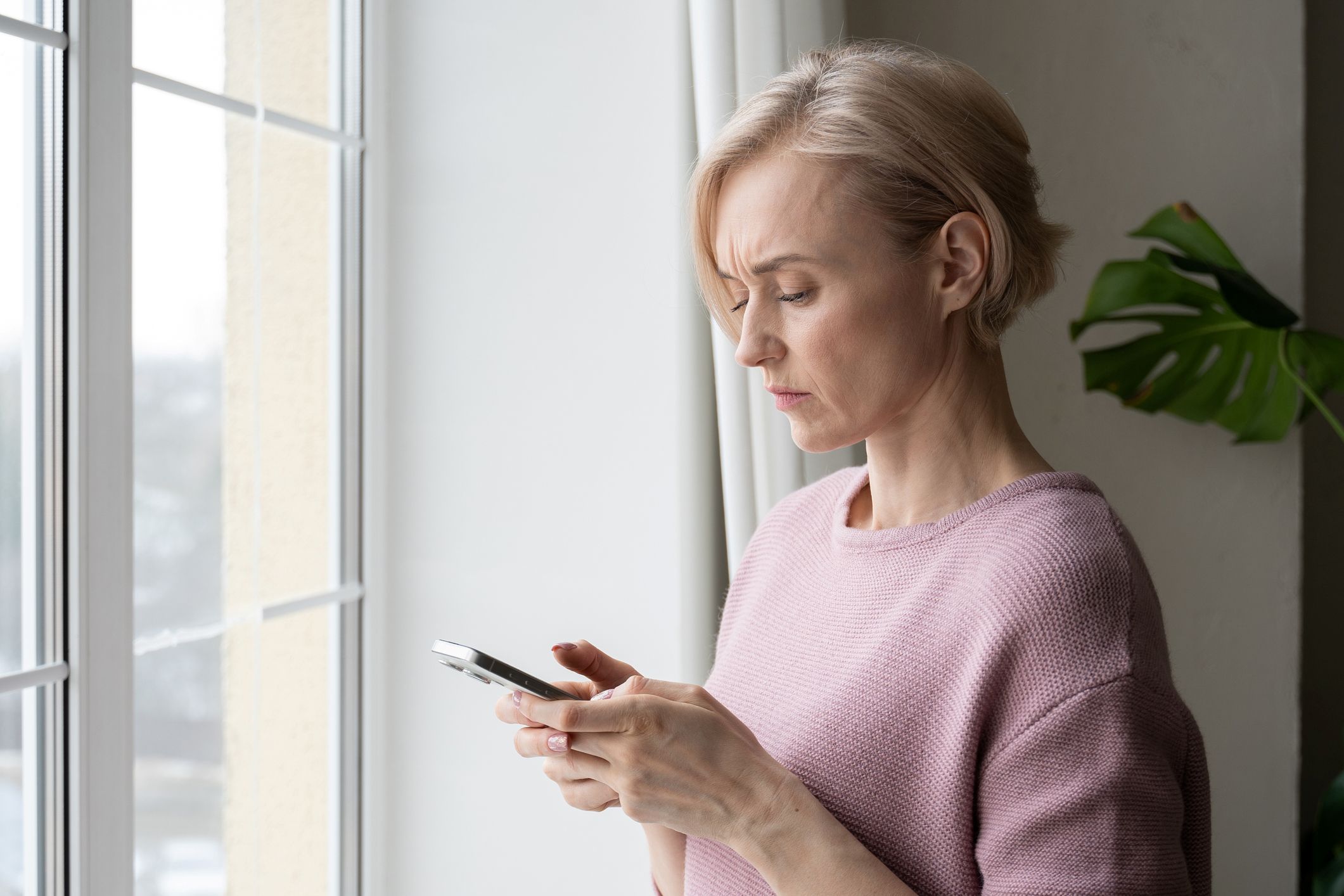 A blond female standing next to the window and looking at her phone